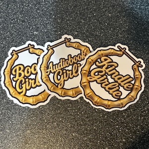 May include: Three white stickers with gold text and bamboo-style hoop designs. The stickers read "Book Girl", "Audiobook Girl", and "Kindle Girl" in a stylish script font. The stickers are on a dark gray surface.