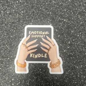 Pode incluir: Um adesivo com uma ilustra&ccedil;&atilde;o de desenho animado de m&atilde;os segurando um Kindle. O Kindle exibe as palavras "EMOTIONAL SUPPORT KINDLE". As m&atilde;os s&atilde;o adornadas com an&eacute;is e pulseiras douradas e t&ecirc;m unhas longas e pontudas.