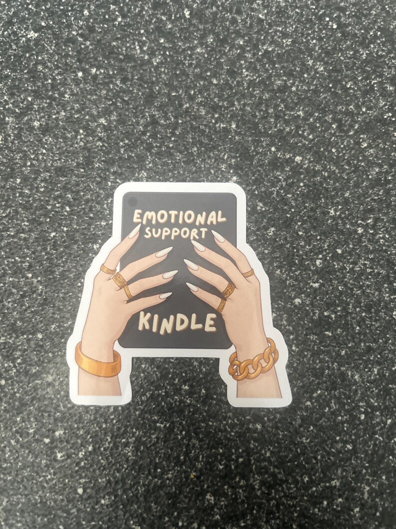 Pode incluir: Um adesivo com uma ilustra&ccedil;&atilde;o de desenho animado de m&atilde;os segurando um Kindle. O Kindle exibe as palavras "EMOTIONAL SUPPORT KINDLE". As m&atilde;os s&atilde;o adornadas com an&eacute;is e pulseiras douradas e t&ecirc;m unhas longas e pontudas.