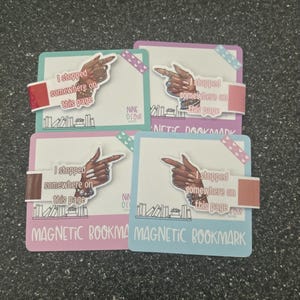 May include: Four magnetic bookmarks with a rectangular design. Each bookmark features a hand pointing towards the text "I stopped somewhere on this page." The text "MAGNETIC BOOKMARK" is at the bottom. The bookmarks are in pink, blue, purple, and teal.