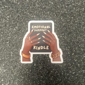 Pode incluir: Um autocolante com uma m&atilde;o de pele escura segurando um ret&acirc;ngulo preto com as palavras "EMOTIONAL SUPPORT KINDLE" em branco. As m&atilde;os s&atilde;o adornadas com an&eacute;is e pulseiras douradas. O autocolante est&aacute; sobre uma superf&iacute;cie cinza salpicada.