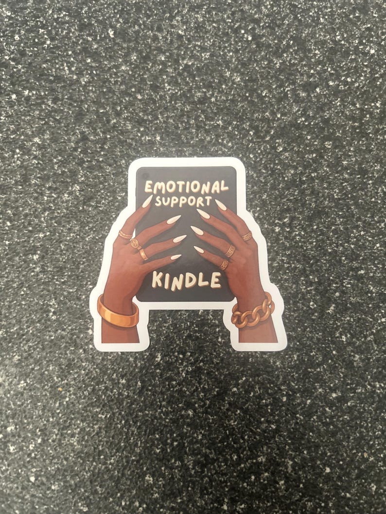 Pode incluir: Um autocolante com uma m&atilde;o de pele escura segurando um ret&acirc;ngulo preto com as palavras "EMOTIONAL SUPPORT KINDLE" em branco. As m&atilde;os s&atilde;o adornadas com an&eacute;is e pulseiras douradas. O autocolante est&aacute; sobre uma superf&iacute;cie cinza salpicada.