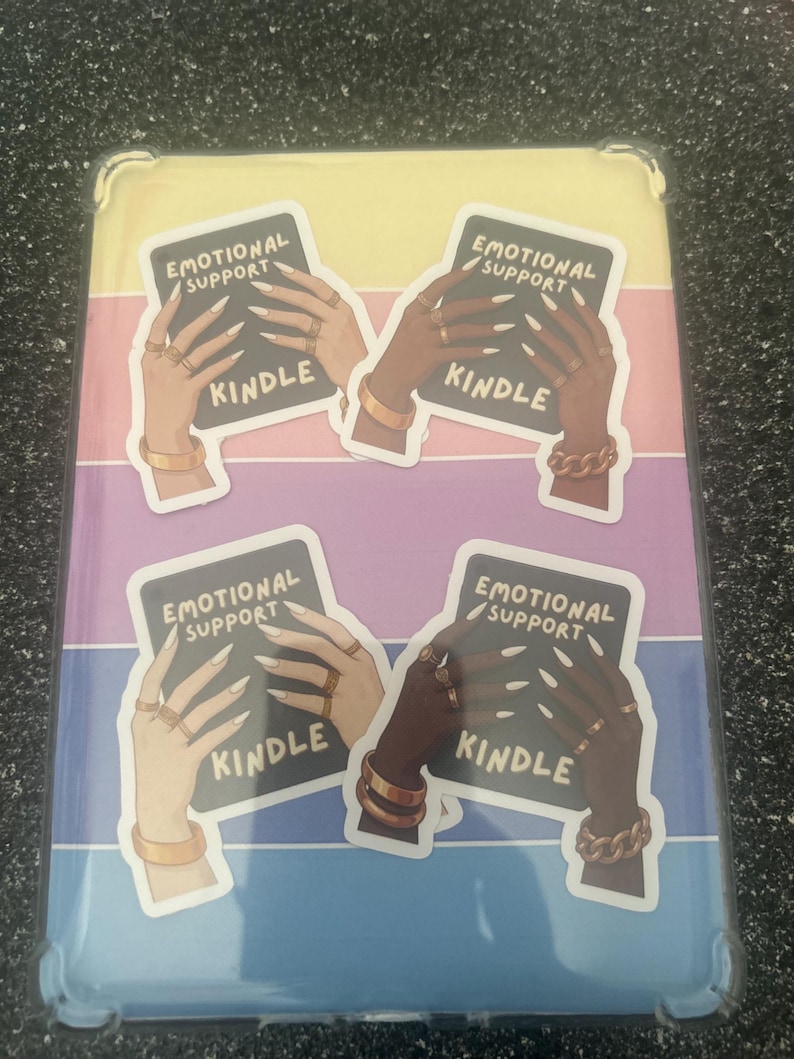 Pode incluir: Capa Kindle com um fundo listrado colorido. A capa apresenta quatro adesivos com o texto "EMOTIONAL SUPPORT" e "KINDLE". Os adesivos retratam m&atilde;os adornadas com joias de ouro.