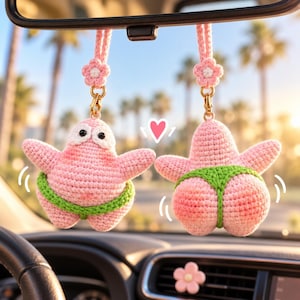 Peut inclure: Deux figurines d'étoiles de mer crochetées en rose suspendues au rétroviseur d'une voiture. Chaque figurine porte un string vert et est attachée à une sangle en fleur crochetée rose avec un fermoir doré. Un petit cœur rose est dessiné entre elles.