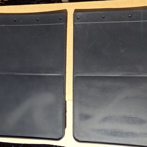 May include: Two black plastic mud flaps with a rectangular shape and a smaller rectangular section at the bottom. The flaps have multiple holes along the top edge.