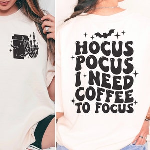 以下が含まれることがあります： 黒いコーヒーカップと中指を立てたスケルトンの手のグラフィックが描かれた白いTシャツ。背面には「Hocus Pocus I Need Coffee To Focus」の文字が書かれている。