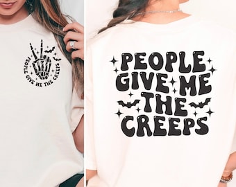Può includere: Una maglietta bianca con una mano scheletrica nera che fa il segno della pace e il testo "People give me the creeps" sul davanti. La parte posteriore della maglietta ha lo stesso testo in un carattere più grande con stelle e pipistrelli.