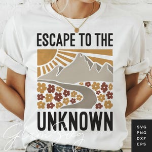 Puede incluir: Camiseta blanca con el texto negro "ESCAPE TO THE UNKNOWN". El diseño presenta una cordillera, un sol y flores en tonos marrón y dorado. Se ven un collar y una pulsera.