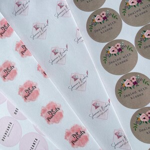 May include: Assortment of round stickers with various designs. Some stickers feature a pink watercolor design with the text "Pup Pawtraits" and a logo with the text "Shereens Bakes" in Arabic script. Other stickers feature a floral design with the text "Sealed with kisses" on a brown background.