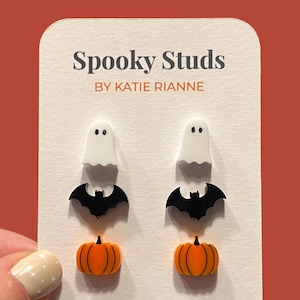 May include: A set of three pairs of Halloween-themed earrings. Each pair features a different design: a ghost, a bat, and a pumpkin. The earrings are made of white, black, and orange plastic.