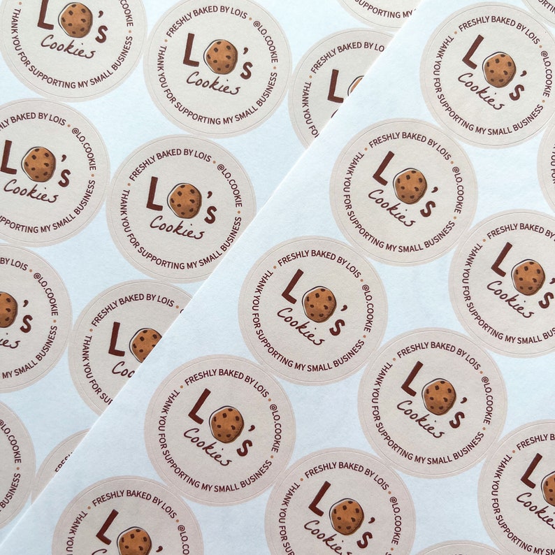 May include: Close-up of several round, white stickers with brown text and a graphic of a chocolate chip cookie. The text on the stickers reads "Lo's Cookies" and "Freshly Baked by Lois @lo.cookie. Thank you for supporting my small business."