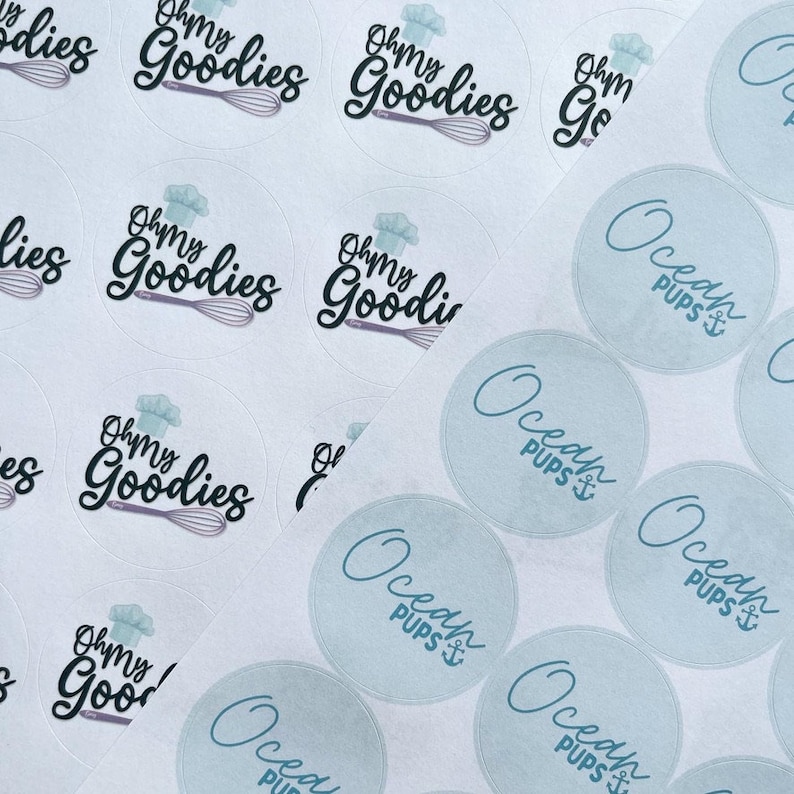 May include: A close-up of two sets of round stickers. One set features a white background with a black and purple chef hat and the text "Oh My Goodies". The other set features a light blue background with the text "Ocean Pups" in a blue cursive font with an anchor symbol.