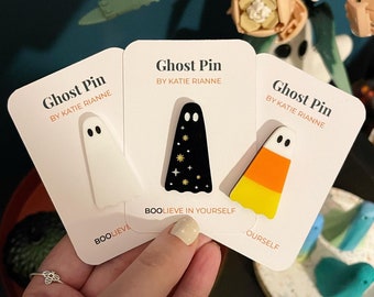 May include: Three white cards with black text and colorful ghost pins. The cards read "Ghost Pin by Katie Rianne". The pins are a white ghost, a black ghost with gold stars, and a candy corn ghost.