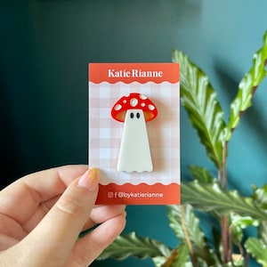 Könnte beinhalten: Eine weiße, geisterförmige Anstecknadel mit einem roten und weißen Pilzkopf. Die Nadel befindet sich auf einem weißen und rosafarbenen Gingham-Hintergrund mit dem Text "Katie Rianne" und "@bykatierianne".