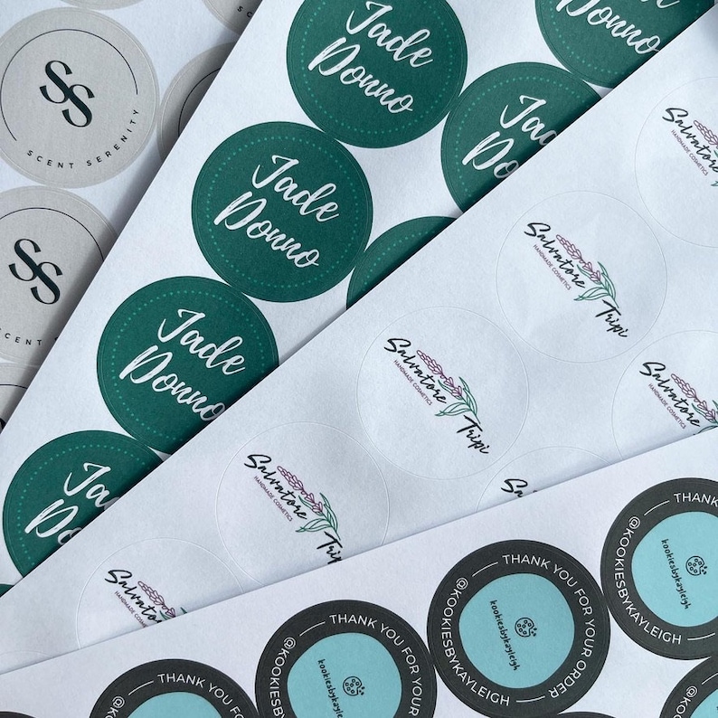 May include: Assortment of round stickers with various designs. Some stickers feature the text "Jade Donno" in green lettering on a green background. Other stickers feature a floral design with the text "Salvatore Tripi" in black lettering.  A few stickers feature the text "Scent Serenity" in black lettering on a beige background.  The stickers are arranged on a white surface.