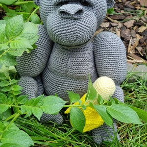 May include: A grey crocheted gorilla toy holding a yellow crocheted banana. The toy is sitting in a bed of green grass.