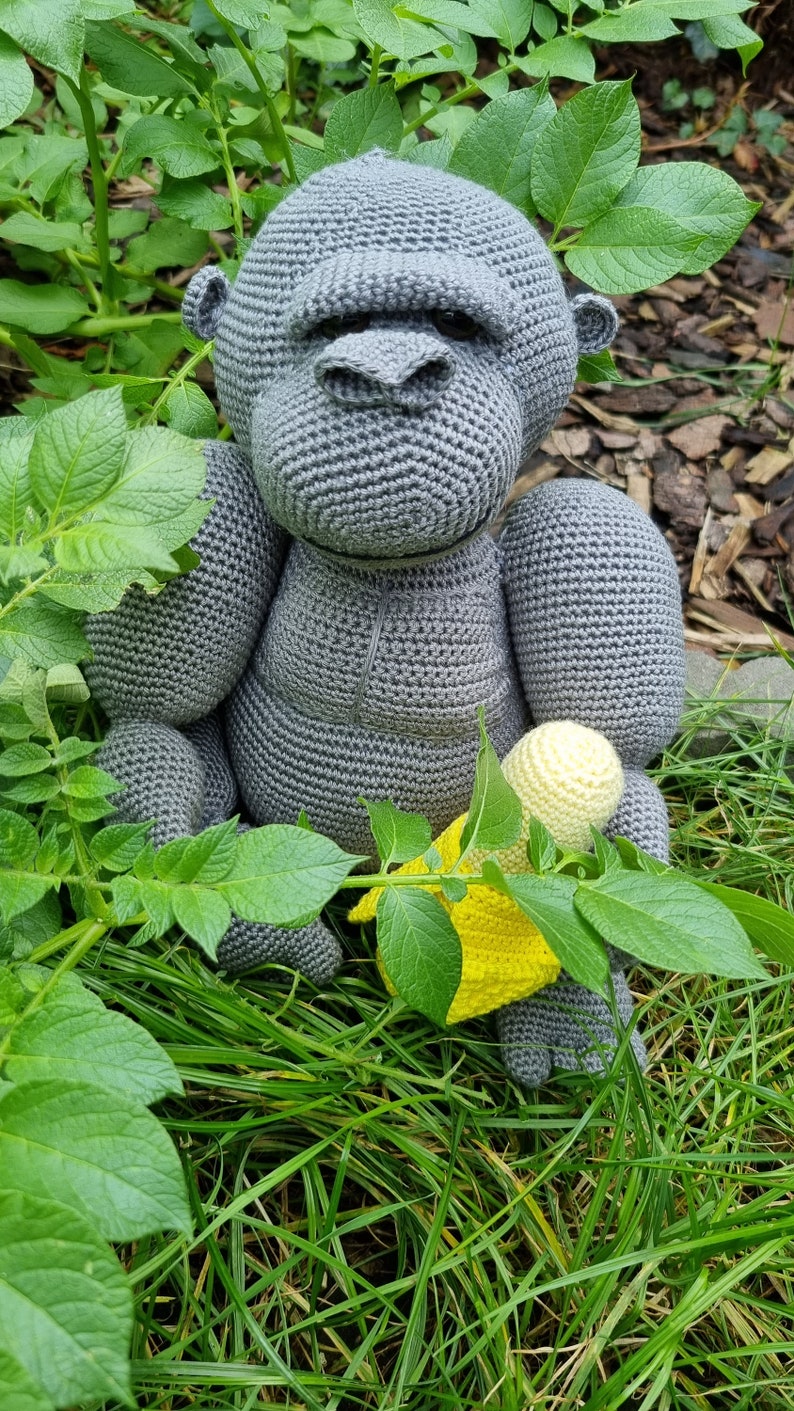 May include: A grey crocheted gorilla toy holding a yellow crocheted banana. The toy is sitting in a bed of green grass.