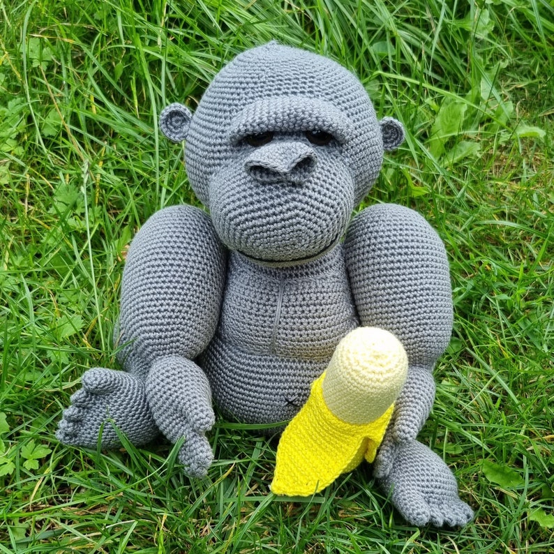 May include: A grey crocheted gorilla toy holding a yellow crocheted banana. The gorilla has a serious expression and is sitting on a bed of green grass.