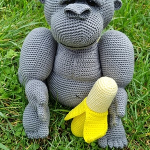 May include: A grey crocheted gorilla toy holding a yellow crocheted banana toy. The toys are sitting on a bed of green grass.