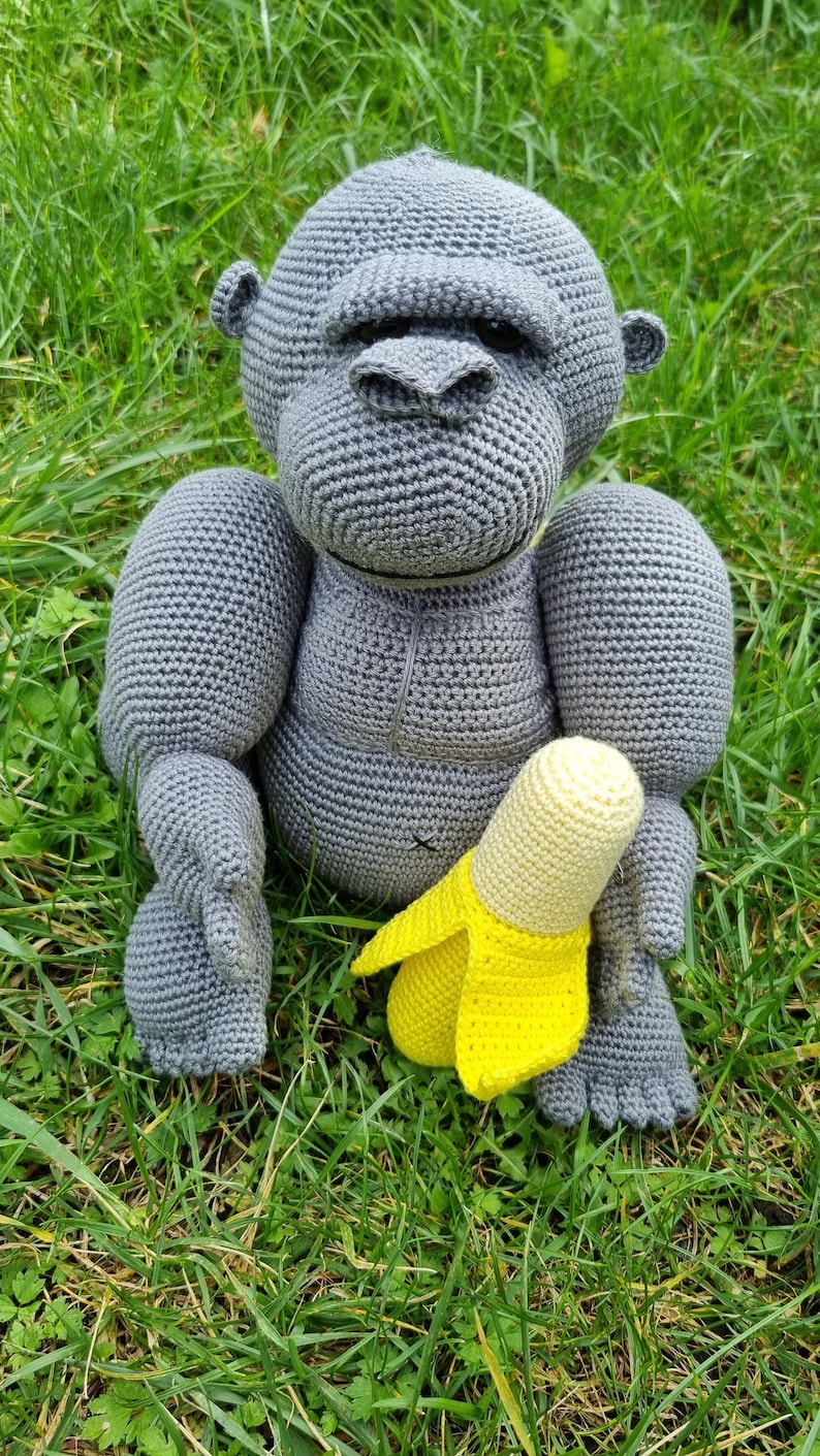 May include: A grey crocheted gorilla toy holding a yellow crocheted banana toy. The toys are sitting on a bed of green grass.