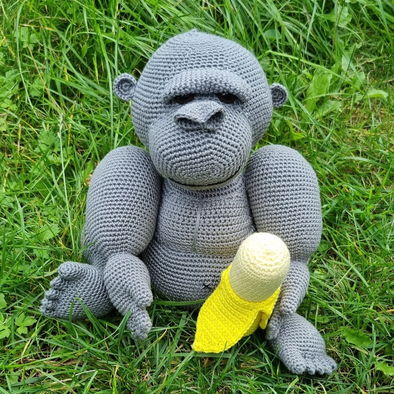 May include: A grey crocheted gorilla toy holding a yellow crocheted banana. The gorilla has a serious expression and is sitting on a bed of green grass.