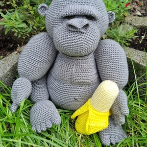 May include: A grey crocheted gorilla toy sitting in green grass holding a yellow crocheted banana.