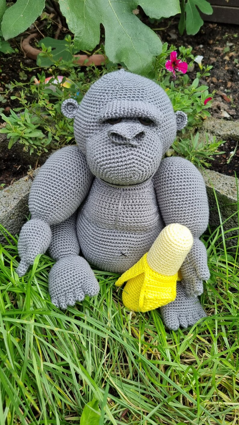 May include: A grey crocheted gorilla toy sitting in green grass holding a yellow crocheted banana.