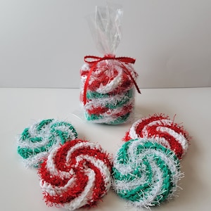 May include: A collection of handmade, spiral-patterned scrubbies in red, white, and green, resembling peppermint candies. Some are packaged in a clear cellophane bag tied with a red ribbon. These round scrubbies are ideal for kitchen or bath use.
