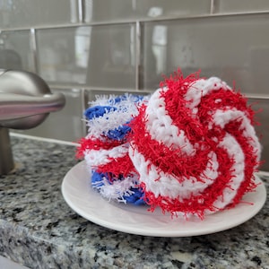 May include: A stack of crocheted dishcloths in red, white, and blue colors. The dishcloths are made with a fluffy yarn and are arranged in a spiral pattern.