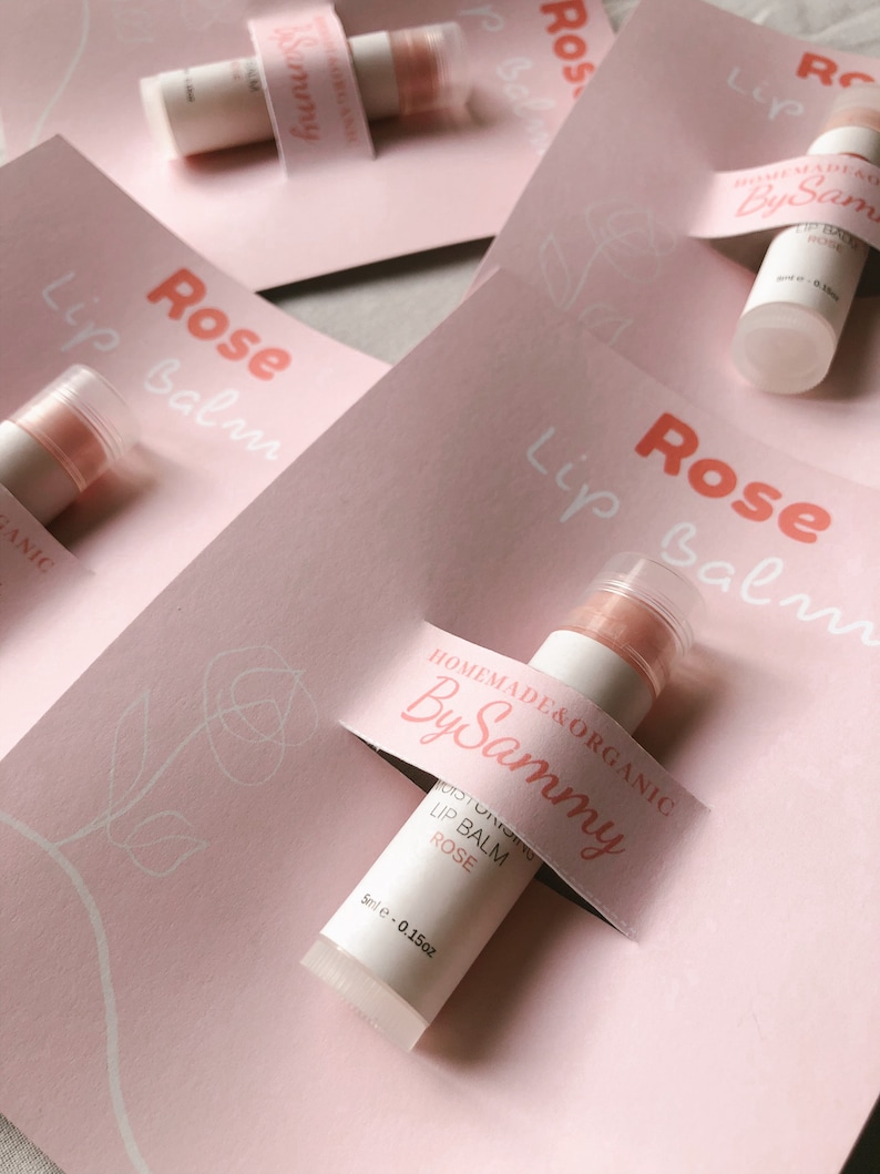 May include: A pink and white label with the text "HOMEMADE & ORGANIC By Sammy" is attached to a clear lip balm tube with a pink balm inside. The label also says "MOISTURIZING LIP BALM ROSE 5ml e-0.15oz".