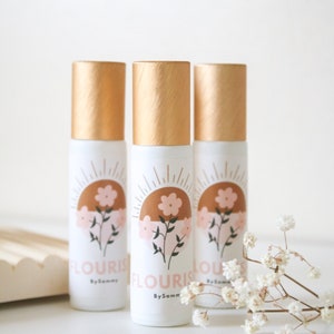 以下が含まれることがあります： ゴールドの蓋と「Flourish by Sammy」と書かれたラベルが付いた白いローラーボトル3本。太陽と花のデザインが施されています。
