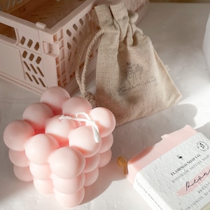 Puede incluir: Una vela rosa con forma de burbujas con una cuerda blanca, una pastilla de jabón rosa con el texto "FLAMINGO SOAP CO. HANDMADE ARTISAN SOAP Peony" y una pequeña bolsa de tela beige con un logotipo de flamenco.