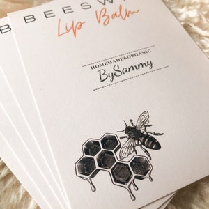 Op de afbeelding: Een stapel witte kaarten met zwarte tekst die "Beeswax Lip Balm Homemade & Organic By Sammy" luidt. De onderste kaart toont een zwart-witte illustratie van een honingraat met een bij.