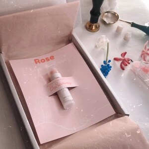 May include: A pink and white box with a pink and white card that says "Rose Lip Balm" and a white tube of lip balm that says "Gimme that moisture" and "By Sammy".