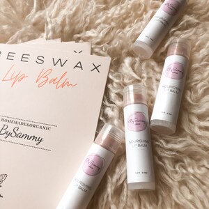 Op de afbeelding: Vier witte tubes lippenbalsem met roze etiketten. De etiketten zeggen "Nourishing Lip Balm" en "By Sammy London".
