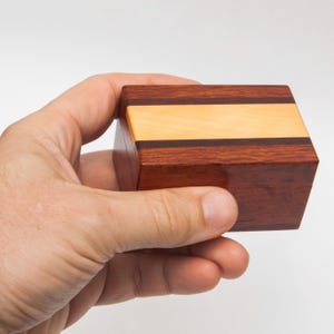 May include: A small rectangular wooden box featuring a horizontal design with a wide light-coloured wood centre stripe flanked by two thin dark wood strips and larger sections of reddish-brown wood on the top and sides of the container.