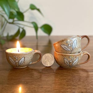May include: Handmade ceramic teacups with leaf and dot designs. One cup holds a lit candle. Two additional cups are stacked. The cups are tan with brown handles and white leaf designs. A quarter is next to the cups.