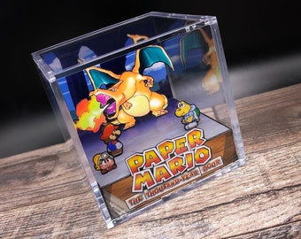 Puede incluir: Una vitrina de acrílico transparente con un diorama 3D de una escena del videojuego Paper Mario: The Thousand-Year Door. El diorama presenta un personaje del juego, una gran criatura naranja similar a un dragón, que escupe fuego, y varios personajes más pequeños. El título del juego está impreso en la base del diorama.