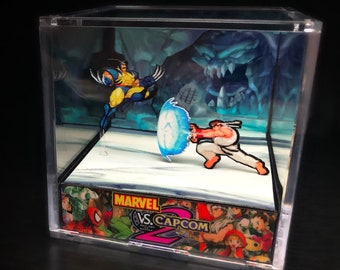 Puede incluir: Un diorama 3D que representa una escena del videojuego Marvel vs. Capcom 2. El diorama presenta a los personajes Wolverine y Ryu en una pose de lucha. El diorama está ambientado en un fondo de un paisaje nevado con una gran criatura amenazante en el fondo. El diorama está encerrado en una caja de acrílico transparente.