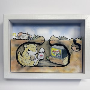 Peut inclure: Une œuvre d'art encadrée représentant une scène de dessin animé avec un personnage de Sabelette tenant une tasse et un biscuit, regardant une télévision montrant une bataille de Pokémon. D'autres personnages Pokémon sont également présents. L'œuvre est située dans un environnement creusé.