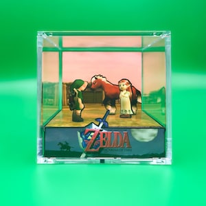Peut inclure: Une vitrine en plastique transparente contenant un diorama de personnages du jeu vidéo "The Legend of Zelda: Ocarina of Time". Le diorama représente Link, un cheval et une jeune fille. La base du diorama porte le logo et le titre du jeu.
