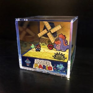 May include: A clear plastic cube display case containing a papercraft diorama. The diorama features a windmill, a purple character, a green character, and a Mario character. The diorama is titled "Paper Mario".