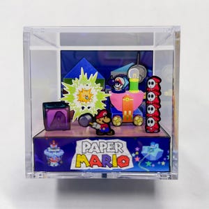 May include: A clear acrylic display case featuring a Paper Mario diorama. The scene includes a Paper Mario figure, a pink vehicle, Shy Guys, and a green explosion effect. The base of the display reads "Paper Mario".