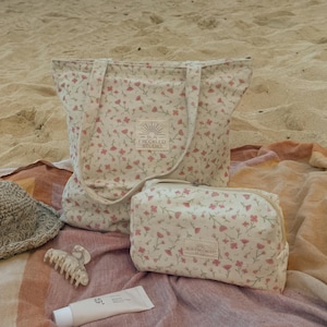 May include: A cream-colored tote bag and a matching rectangular zippered pouch, both featuring a small pink floral pattern and a square label that reads Sun Freckled Studio. Also included are a woven straw hat, a hair claw clip, and a tube of cream.