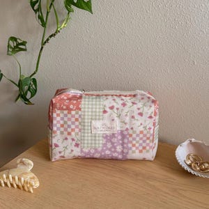 May include: A rectangular fabric pouch featuring a patchwork design of floral and checkered patterns in shades of pink, green, and white. A small rectangular label on the front displays the text SUN FRECKLED STUDIO with a sun icon.