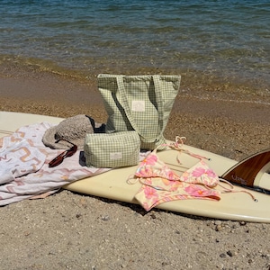 May include: A light green and white gingham tote bag and matching zippered pouch, a beige woven sun hat, brown sunglasses, a white towel with orange patterns, and a pink and yellow floral bikini set arranged on a white surfboard.