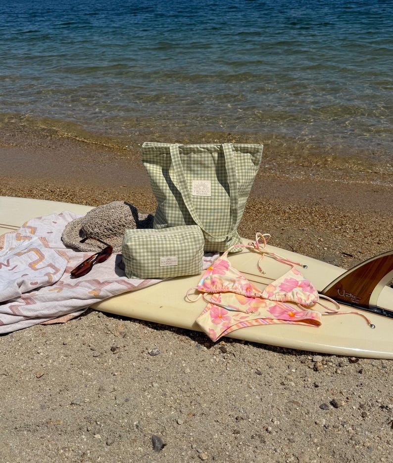 May include: A light green and white gingham tote bag and matching zippered pouch, a beige woven sun hat, brown sunglasses, a white towel with orange patterns, and a pink and yellow floral bikini set arranged on a white surfboard.