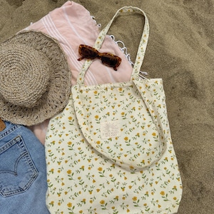 May include: A cream-colored tote bag with a yellow floral pattern, a straw hat, and sunglasses rest on a pink striped towel. The tote bag has a label that reads "FRECKLED STUDIO". A pair of blue denim shorts is also visible.