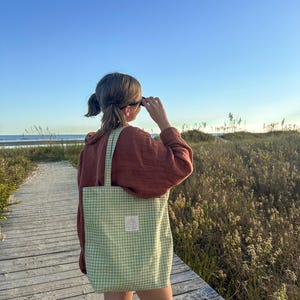 May include: A light green and white gingham patterned tote bag with two shoulder straps and a small rectangular white label sewn onto the front center. The bag is made of fabric and features a consistent checkered pattern throughout the exterior.
