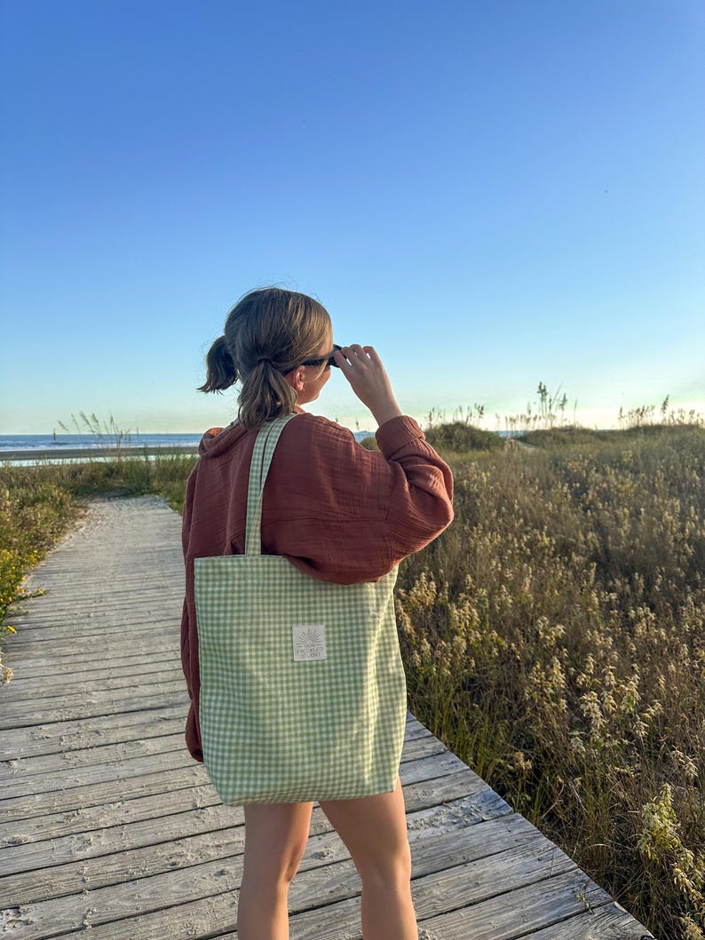 May include: A light green and white gingham patterned tote bag with two shoulder straps and a small rectangular white label sewn onto the front center. The bag is made of fabric and features a consistent checkered pattern throughout the exterior.